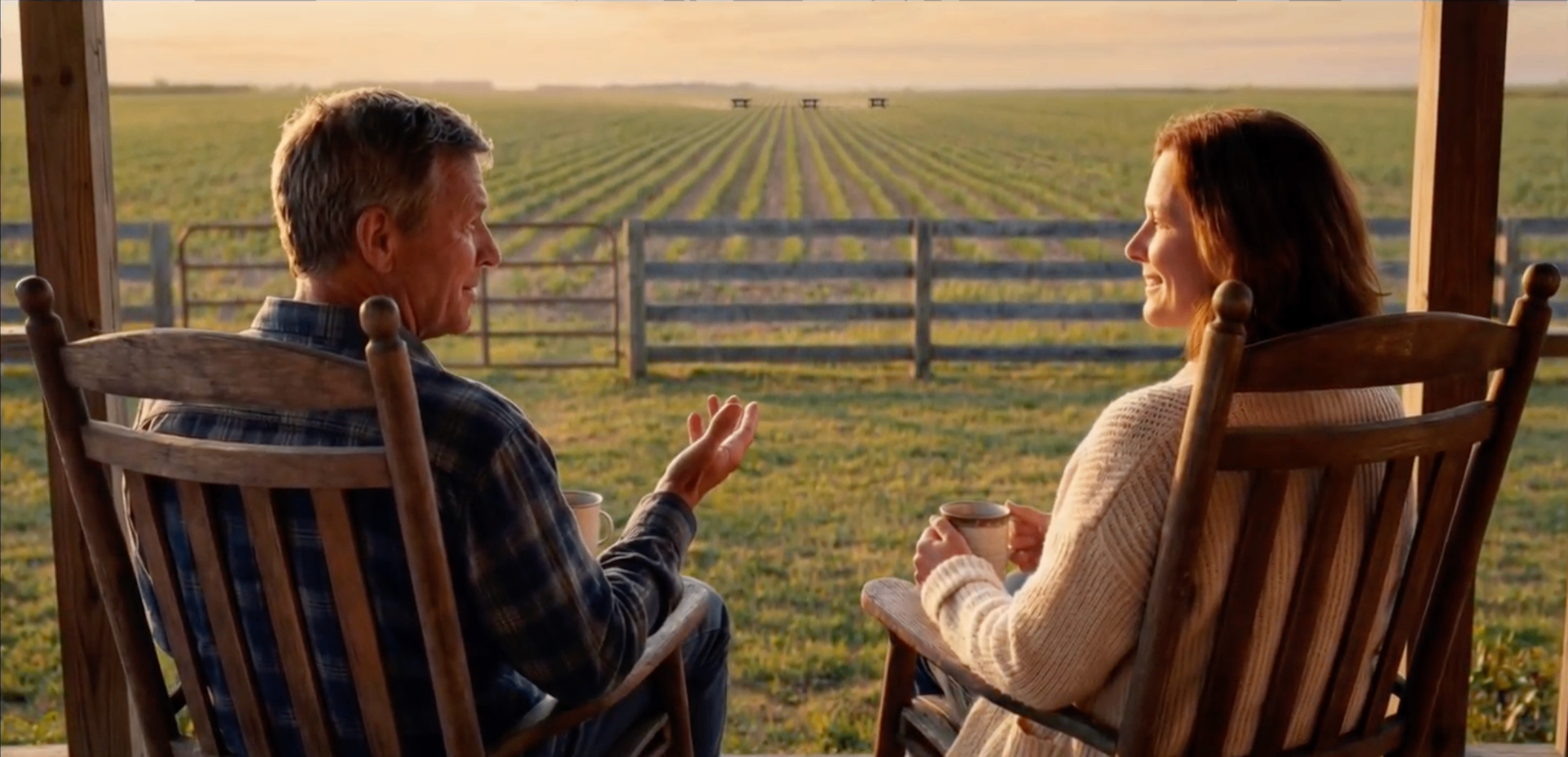 Farmer couple on porch overlooking a field worked by autonomous Solix robots at sunset
